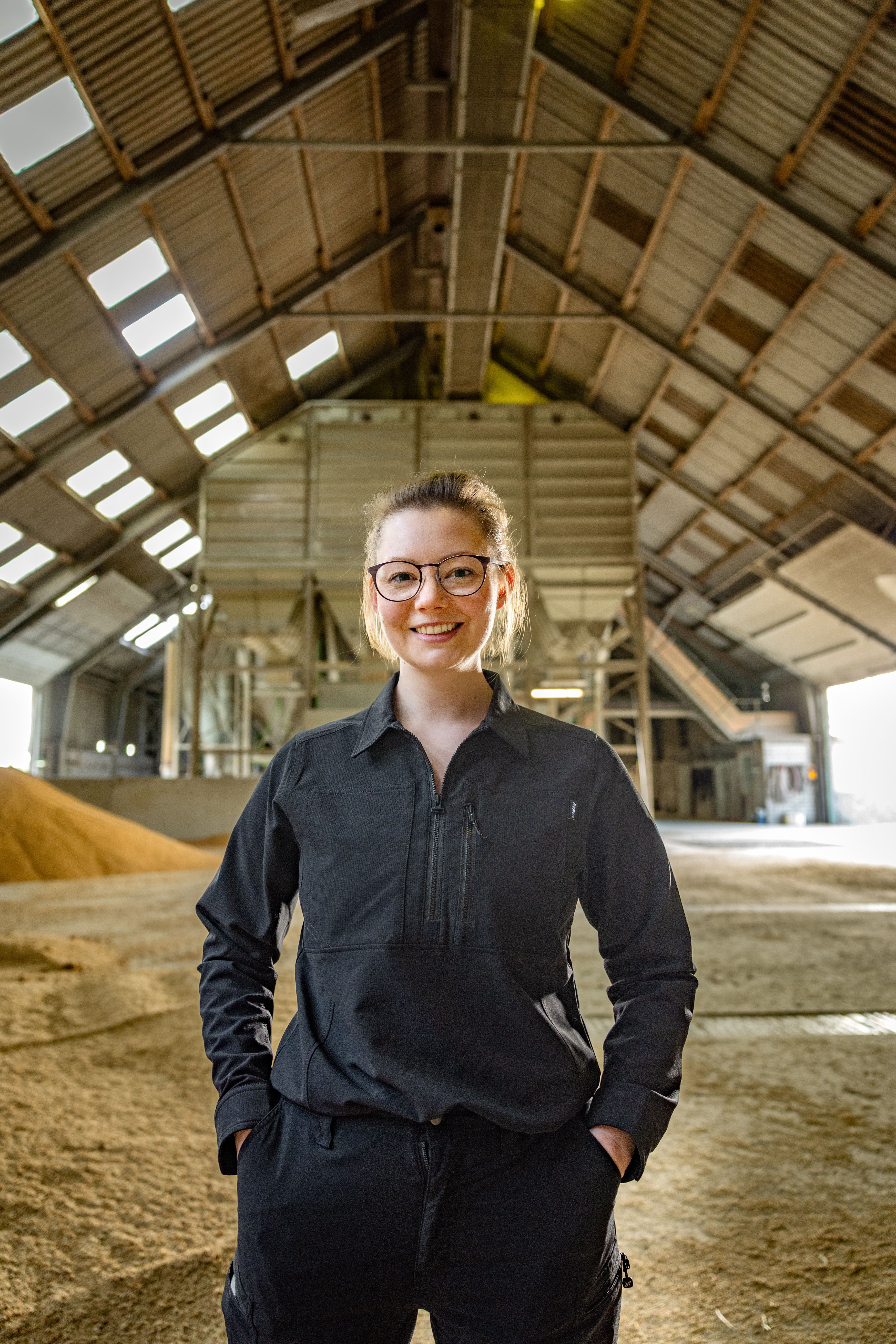 Jenna Struck steht in einer großen, lichtdurchfluteten Halle mit hohem, schrägem Dach. Im Hintergrund sind industrielle Anlagen und Schüttgut zu sehen. Sie trägt eine schwarze, langärmelige Arbeitsjacke und eine dunkle Hose, die Hände in den Hosentaschen. Das Licht fällt durch die Fenster im Dach und erzeugt helle Flächen auf dem Boden. Jenna Struck steht in einer großen, lichtdurchfluteten Halle mit hohem, schrägem Dach. Im Hintergrund sind industrielle Anlagen und Schüttgut zu sehen. Sie trägt eine schwarze, langärmelige Arbeitsjacke und eine dunkle Hose, die Hände in den Hosentaschen. Das Licht fällt durch die Fenster im Dach und erzeugt helle Flächen auf dem Boden.