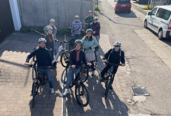 Pfalz Mehrere Personen mit Fahrradhelmen stehen mit Fahrrädern auf einem gepflasterten Hof. Die Gruppe blickt nach oben in Richtung Kamera. Im Hintergrund sind ein grauer Zaun, ein geparkter weißer Transporter und ein rotes Auto auf einer asphaltierten Straße zu sehen. Die Szene wirkt sonnig, die Schatten sind klar erkennbar.